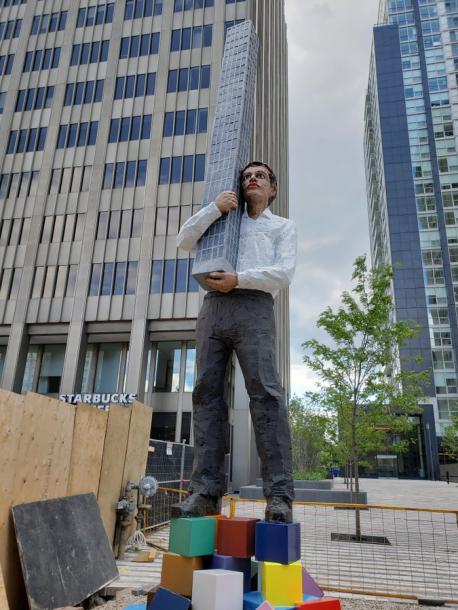 One pre-Pandemic art piece that has stirred controversy since it was installed is a 25-foot-tall, expressionless man holding a skyscraper in his arms atop a pile of colourful cubes, in Toronto., and has been nicknamed the 